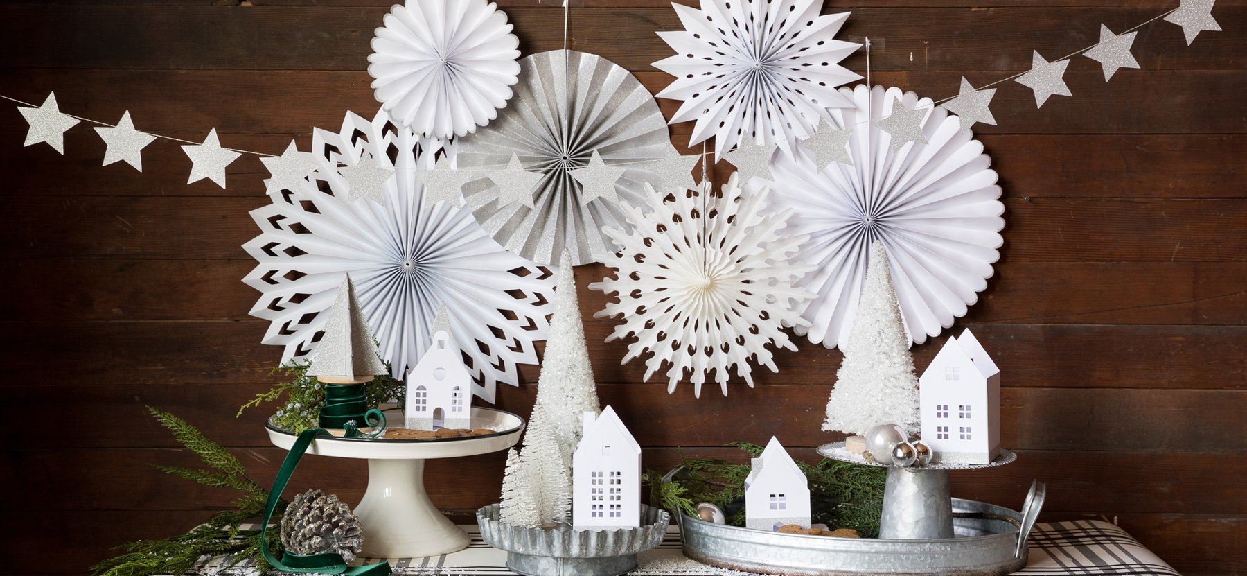 Winter White Christmas holiday home table set up  with My minds eyes' White Glitter Stars Banner in front of winter white party fans as wall decoration, white christmas brush trees with white house candle holders, and silver ornaments on the galvanized steel tray and cake stands, with green ribbon and green plants decorated for a white winter cozy holiday celebration