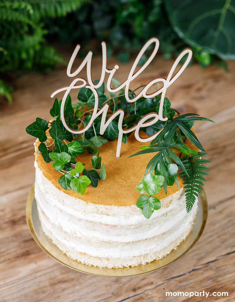 Cake decorated with Party Deco Wild One Wooden Cake Topper and greens on top. With This modern designed, 8.6 inches height, "Wild One" Script letter wooden topper, on a wooden table, ready for a modern safari themed 1st birthday party!