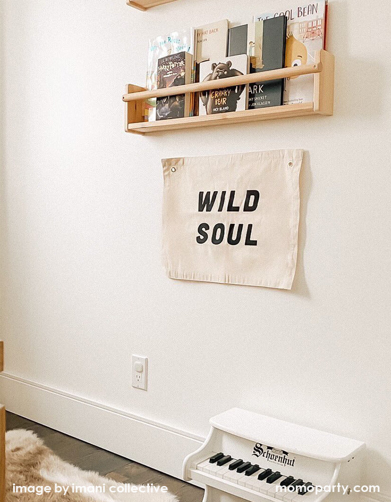 Room room decor with Imani Collective Wild Soul Banner. Nature canvas with black screen print text of Wild Soul, hanging under a kids wooden book shelf, above a white piano toy 