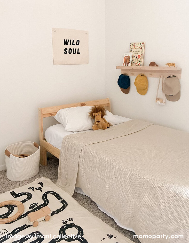 A Modern california style kid's room decorated with Imani Collective Wild Soul Banner. Nature canvas with black screen print text of Wild Soul, hanging on the top of  kids wooden framed bed, a book shelf with few hats hanging on it, books and toy on the top, a road floor map on top of a light color rug. 