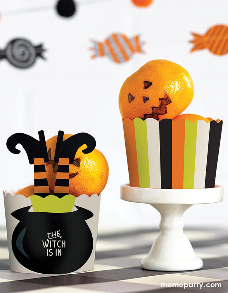 Table top with tangerines in the My Minds Eye - WITCH IS IN FOOD CUPS. one with Witch Leg Stakes with "the witch is in" text in the Cauldron. another cups in a mint, orange, white and black stripe design with tangerine drawed as a jack-o-lantern.  These fun halloween themed  baking cups are perfect for baking Halloween cupcakes right in the oven.