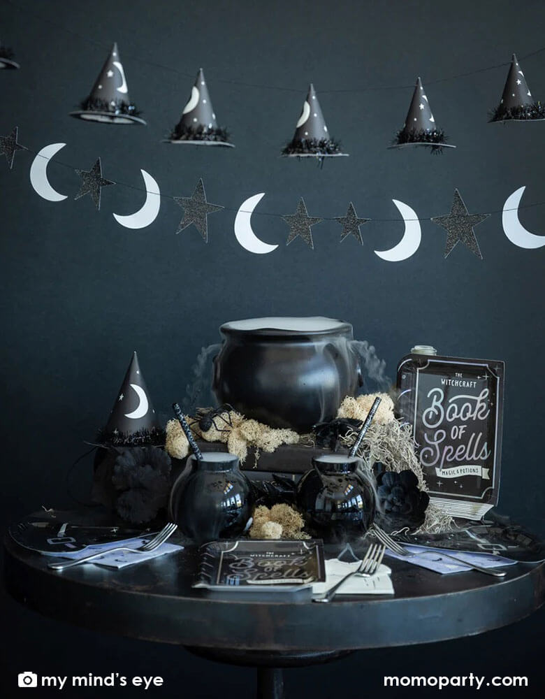 A Halloween witch themed party table set up with cauldron of witch brew and witch themed tableware including My Mind's eye book of spell shaped plates, witch hat shaped plates, witch candle napkins and witch hats, with moon and star and witch hat shaped banner hung on above, all creates a spooky vibe for a frightening Halloween Eve