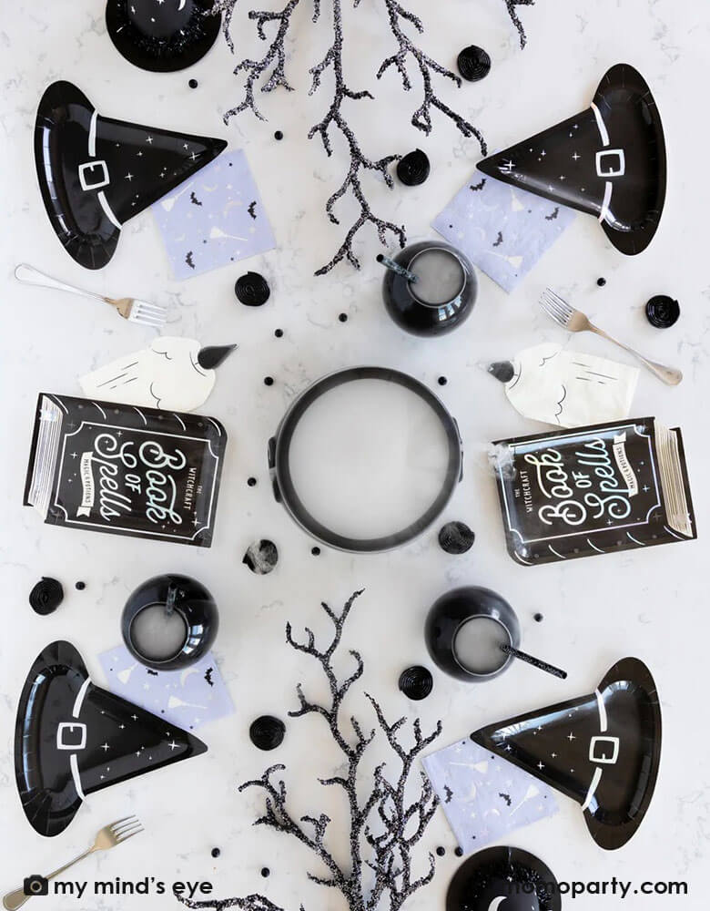 A witch themed Halloween party tablescape featuring My Mind's Eye Witching hour tableware including witch hat shaped plates, book of spell shaped plate, witch candle napkins, witching hour lilac small napkins with crawling branches in black and witch brew in cauldrons, creating a spooky vibe for a frightening Halloween Eve bash!