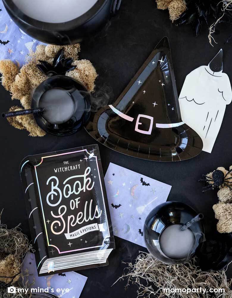 A Halloween witch themed party table filled with witch inspired tableware including My Mind's eye witch hat shaped plates, book of spells shaped plates, lilc witching hour small napkins and cauldrons for witch brew