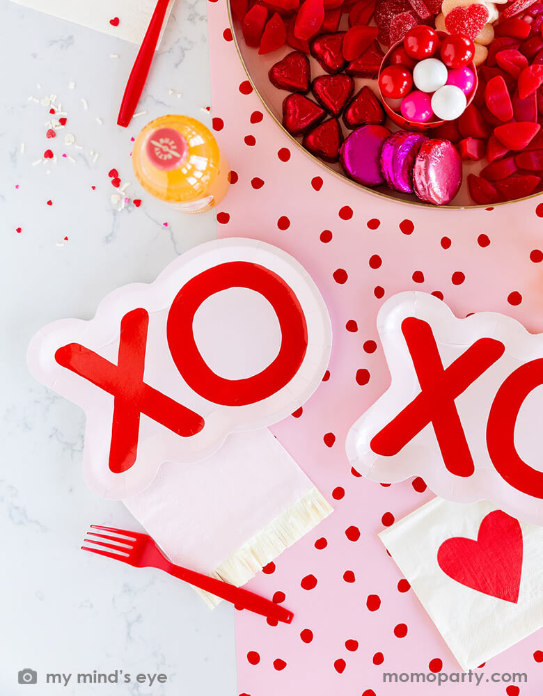 A Valentine's Day table set up featuring Momo Party's XO party plates, red dotted pink table runner, and heart napkins by My Mind's Eye, with the centerpiece of a plate full of Valentine's Day chocolates in heart shaped and red and pink wrappers, all makes a sweet tablescape for a Valentine's Day or a girl's Galentine's Day party.