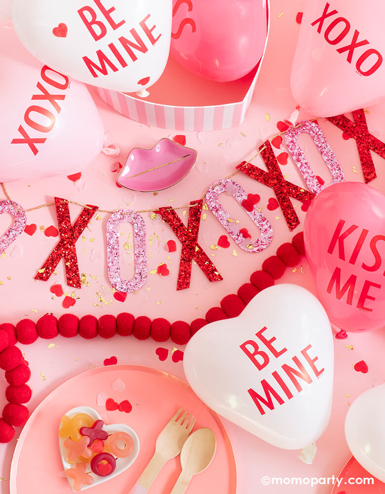 Valentine day table decoration with lots of mini conversation hearts balloons around StudioPepe XOXO Artisan Red & Pink glitter fabric Banner, kiss me heart shaped confetti , Oh happy day pretty in pink plates, and red ball felt garland, xoxo jelly candies in a heart shape plates for a sweet valentine, galentine party