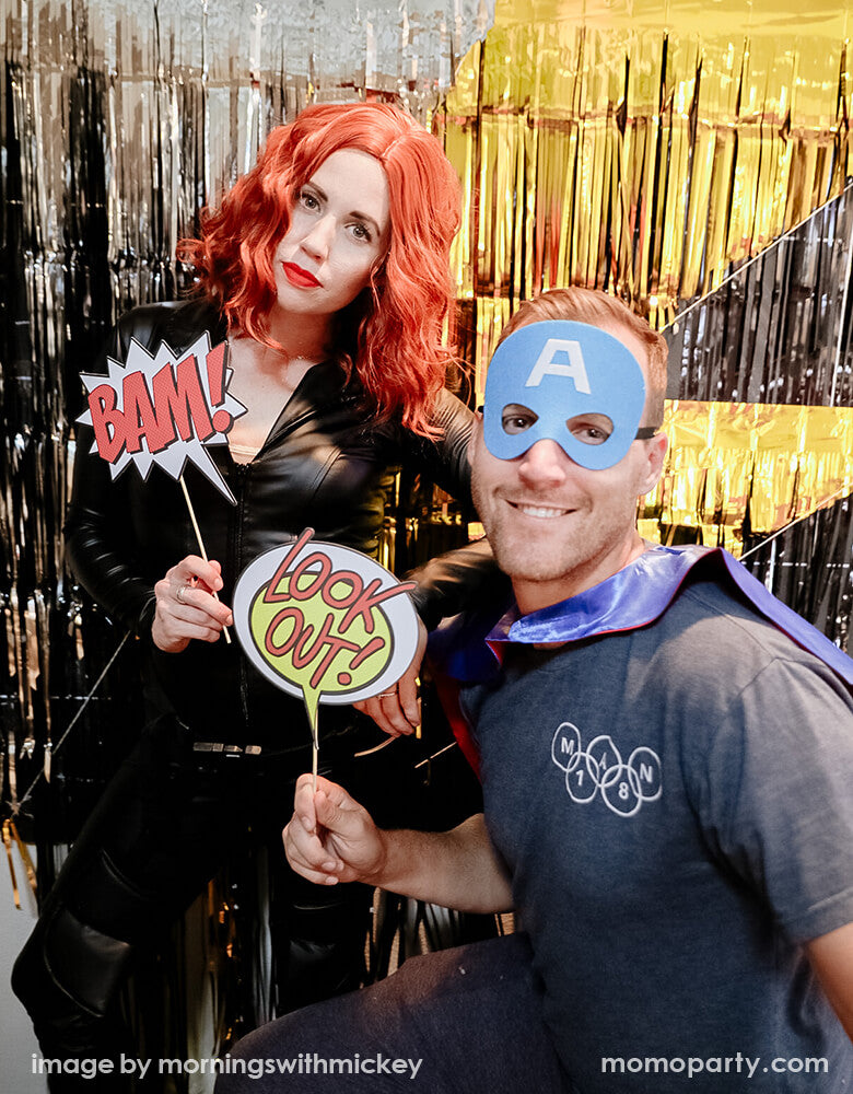 a couple wearing a superhero and catwoman costume, holding My Minds Eye - Paper Love Comic Pop Photo Props, taking photo in front of a lighting backdrop curtain