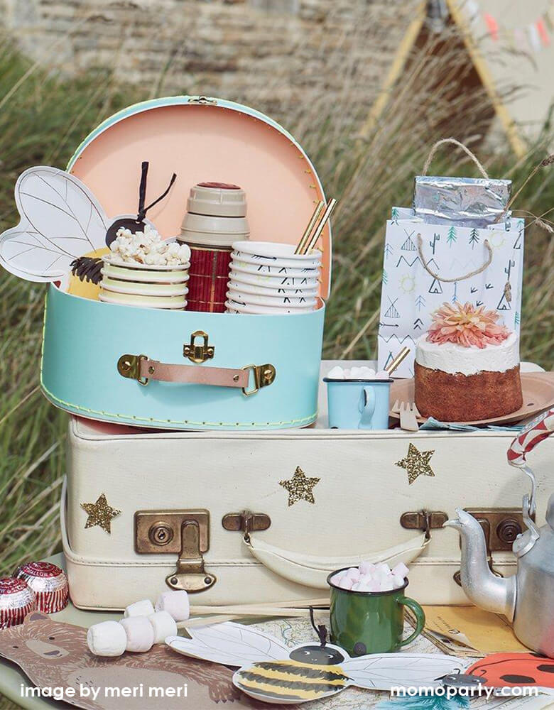 A small table filled with Meri Meri Ladybug Napkins, Bee Plates, Brown Bear plate, a green vintage cups with hot chocolate and mini marshmallows. Let's Explore cups with popcorns, bee plates and a water bottle inside a vintage pale blue Suitcase, Let's Explore party bag with a small cake on a large beige colored vintage style suitcase, This fun modern set up is perfect for a outdoor woodland camping party in summer, camping themed birthday party, summer activities, autumn woodland themed birthday party